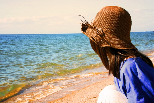 辛い時こそ笑えと言われても笑えないと浜辺で海を眺めている女性の画像